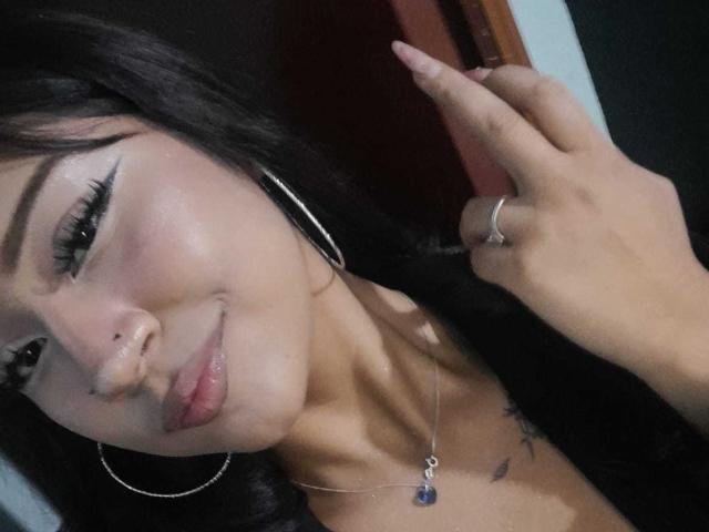 BonnieBodn met donker haar, oorringen, neusring, eyeliner, lichtroze huid, lichte glimlach, hand omhoog, ring, ketting met een paarse steen, kleine tatoeages op de borst..