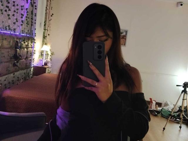BonnieBodn, avec de longs cheveux bruns, portant un haut noir à une épaule, prend un selfie devant un miroir, montrant ses épaules dénudées et tenant un téléphone gris, dans une pièce peu éclairée..