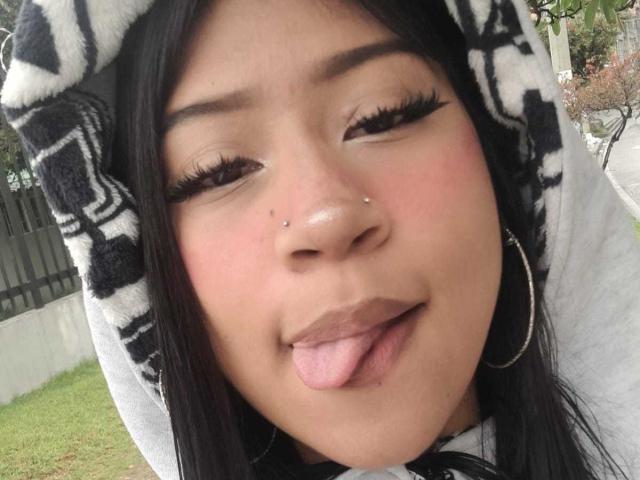 Het gezicht van BonnieBodn, met haar roze tong uitgestoken, zwarte eyeliner, lange wimpers, een neusring, en een zwart-witte hoodie, met grote oorringen..