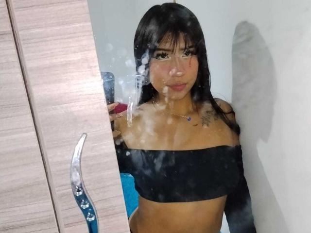 BonnieBodn, avec ses cheveux noirs, portant un haut noir à une épaule, dévoilant ses petites seins et ses tatouages, se prend en selfie dans un miroir avec des traces d'eau..
