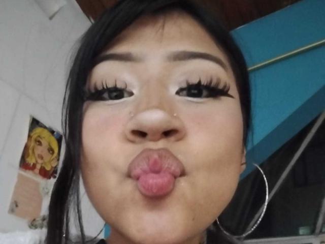 Het verleidelijke gezicht van BonnieBodn, met volle, roze lippen, zwarte eyeliner, lange wimpers en oorringen, maakt een kusgezicht. Aziatische vrouw met donker haar en lichtrode wangen..