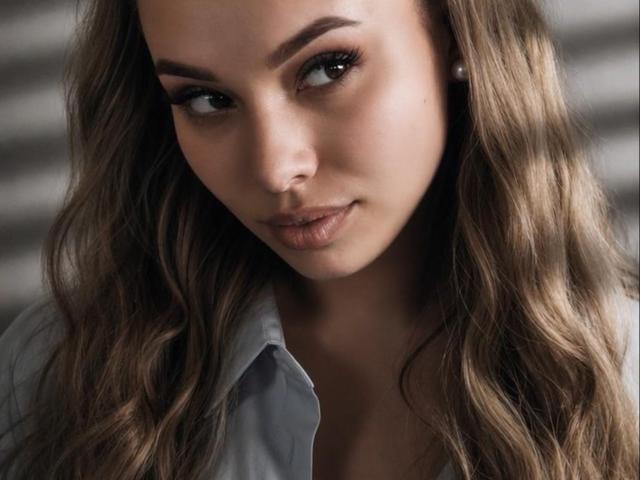 Niferttiti has long, wavy brown hair, light skin, and brown eyes. She's looking slightly to the side with a subtle, sexy smile and full lips. She's wearing a grey shirt.