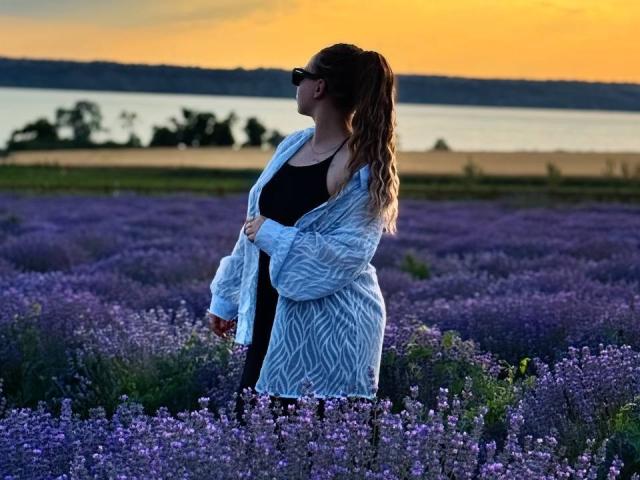 Niferttiti, com cabelos castanhos ondulados, usando óculos de sol, uma blusa preta e um cardigã leve com padrão azul claro, está em pé em um campo de lavanda roxa ao pôr do sol..