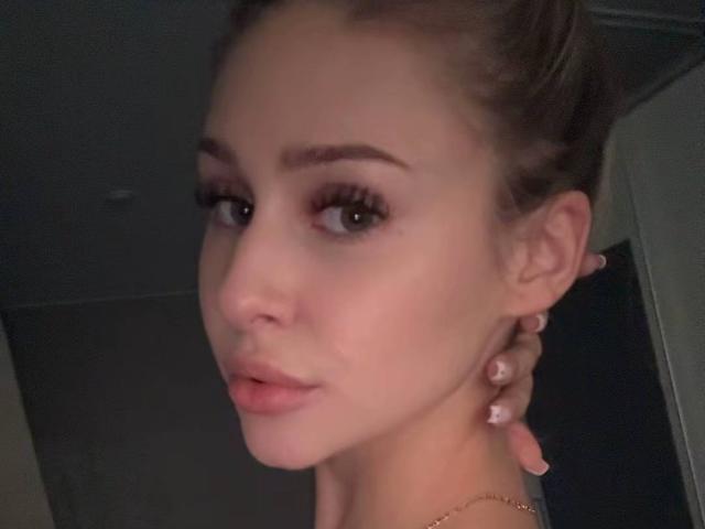 MilagrosEdmisten's face with full lips, light skin, brown eyebrows, long eyelashes, and pink nail polish, holding her neck with a hand, in a dimly lit room.