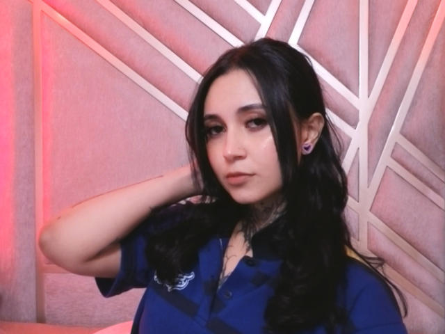GabyMathew has long black hair, wearing a blue shirt, with a hand in her hair, looking serious, purple earrings, and subtle makeup.