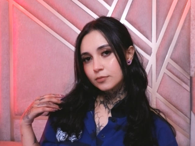 GabyMathew with long black hair, blue shirt, tattoos, purple earrings, holding hair, subtle smile, pink-red lighting, confident look.