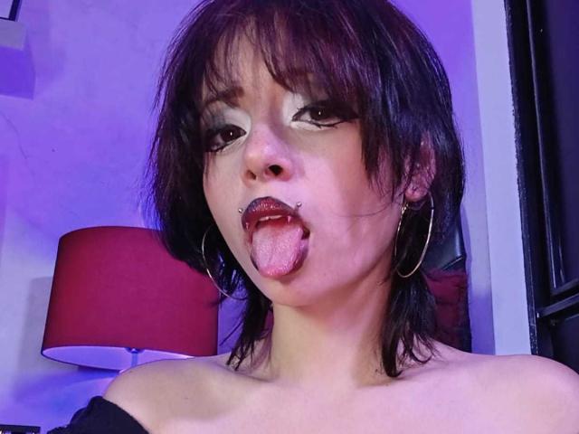 ViolettClay, avec ses cheveux noirs en bataille, ses boucles d'or, et un piercing au nez, tirant la langue avec un entonnoir dans la bouche, portant un haut noir à une épaule..