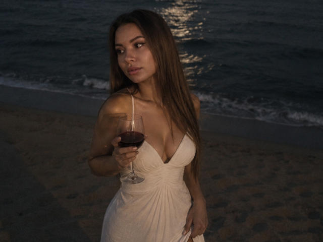 Niferttiti, con cabello largo y castaño, vestida con un vestido blanco, sosteniendo una copa de vino tinto, de pie en una playa al atardecer, mirando hacia un lado con una expresión seria..