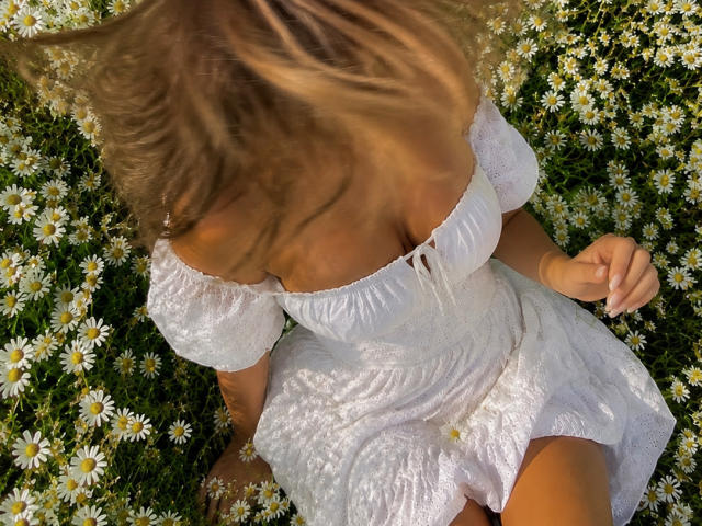 Niferttiti with blonde hair, wearing a white, off-shoulder, lace dress, sitting among daisy flowers, her tits slightly visible, her head down, sunlight filtering through.