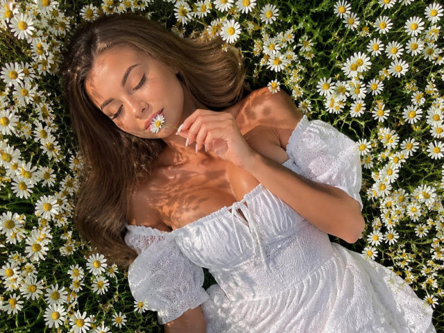 Niferttiti with closed eyes, holding a daisy to her mouth, wearing a white off-shoulder dress, lying in a field of white daisies. Her tits are visible.
