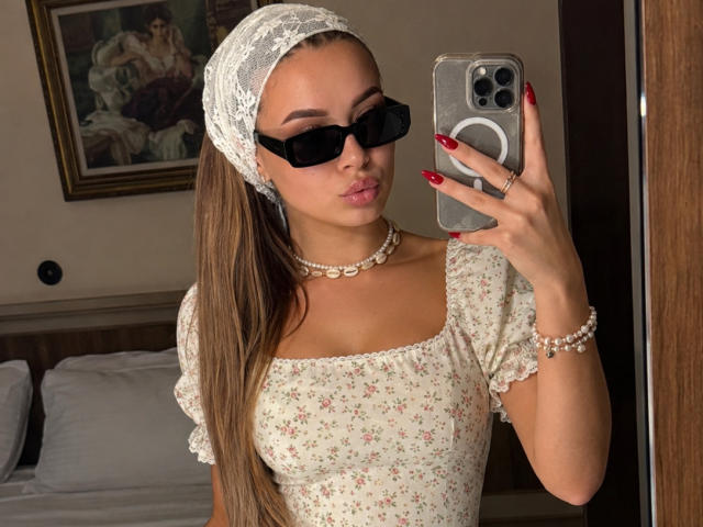 Niferttiti with long brown hair, wearing black sunglasses, white lace headscarf, floral dress, pearl necklace, red nails, holding phone, pouty lips, taking mirror selfie, showing tits.