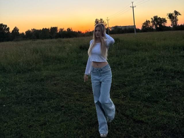 PatriceSorice with long blonde hair, white crop top, blue jeans, and white sandals, standing on grass at sunset, holding her hair, showing her flat stomach and small ass.