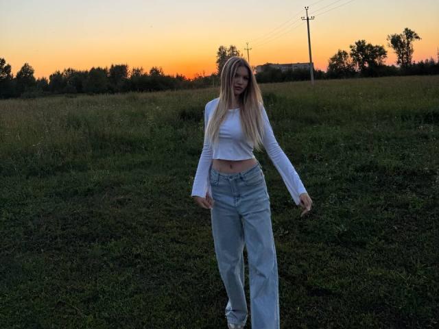 Patrice Sorice, com cabelos loiros longos, uma blusa branca cropped e jeans de cintura alta, está em um campo gramado ao pôr do sol, com o bumbum levemente visível e os braços relaxados..