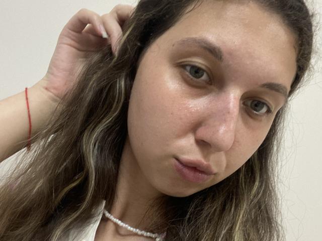 Sanella has a serious expression, slightly puffy face, light brown wavy hair, wearing a white top with a pearl necklace, red bracelet on her right wrist, and looks tired.