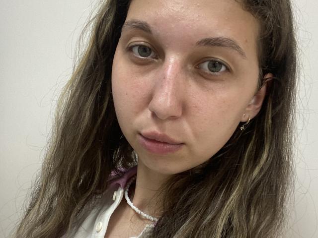 Sanella has a light skin tone, green eyes, and long, wavy brown hair. She's wearing a white shirt with a pink ribbon and a pearl necklace.