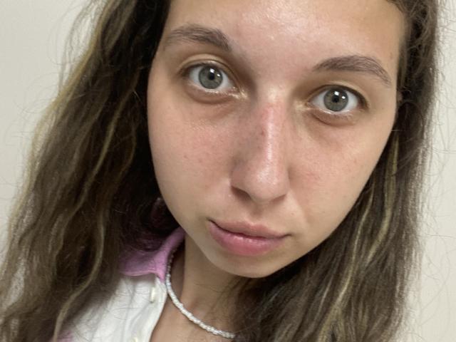 Sanella has pale skin, green eyes, and brown wavy hair. Her face is slightly puffy, with pinkish lips and a subtle nose. She's wearing a white shirt with a pink collar.
