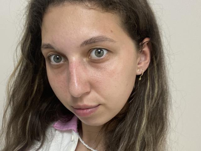 Sanella has light brown, wavy hair, green eyes, and a slight smile. She's wearing a white shirt with a pink collar, a pearl necklace, and gold earrings.