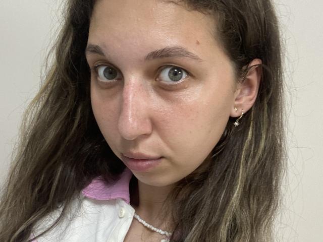 Sanella has a serious face with light green eyes, slightly wavy brown hair, and a small nose. She's wearing a pink and white shirt with a pearl necklace.