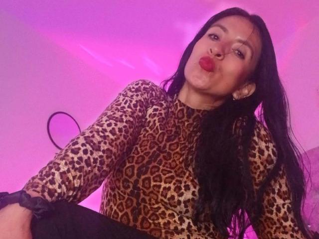 ZarayFeet has long black hair, is wearing a leopard print top, and is puckering her red lips. Her ass is partially visible. Purple lighting highlights her face.