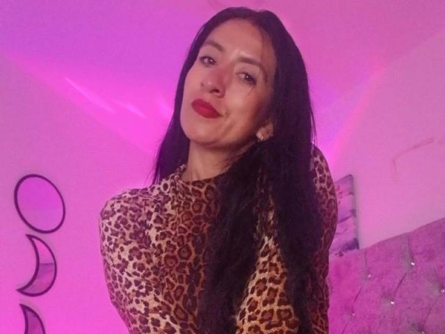 ZarayFeet with long black hair, wearing a leopard print top, pouty red lips, and slightly bent head, in pink-lit room. Boobs visible, no other body parts shown.