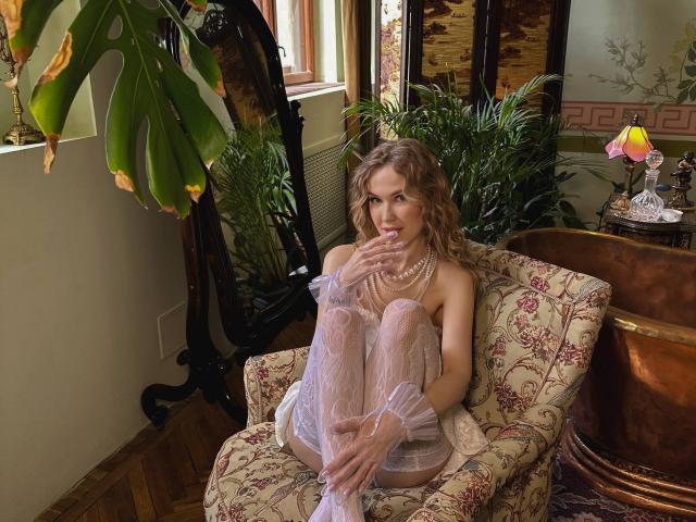 Veronika, avec ses cheveux bruns ondulés, assise sur un fauteuil fleuri, portant une robe blanche transparente, des bas et des gants, touchant ses lèvres, avec une expression pensive, dans une pièce confortable..
