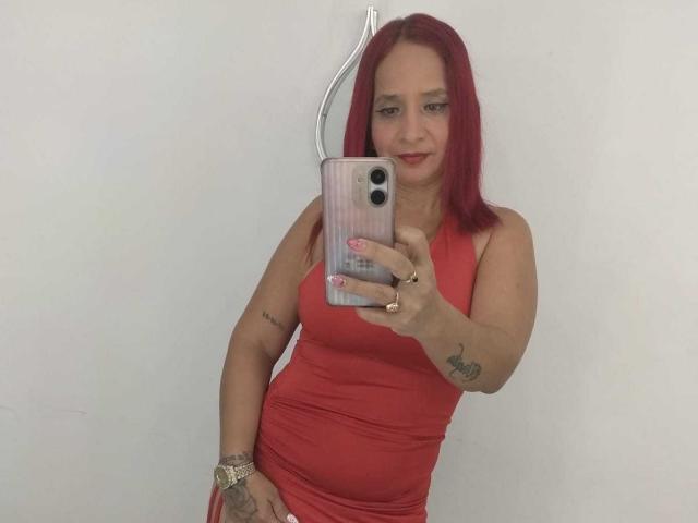 KateMonroe met rood haar, gekleed in een rode jurk, die een selfie maakt met een telefoon. Ze heeft tatoeages op haar arm en hand, en een gouden horloge..