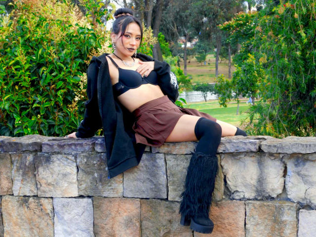 HenutmireRossi with black bra, brown skirt, black cardigan, and thigh-high fringed boots, sitting on stone wall, showing her tits, looking at camera. Greenery in background.