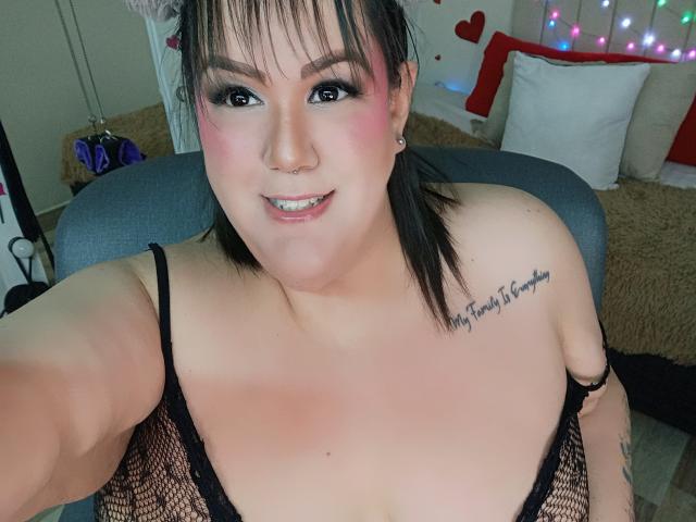 GiiannaMorrys with pale skin, black fishnet top, visible tits, small nose ring, black tattoo on right shoulder, smiling, black and pink hair, colorful lights in background.