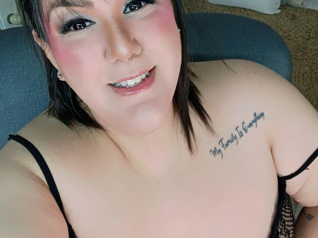 GiiannaMorrys has pink blush, black eyeliner, nose ring, smiling with teeth, black hair, tattoo on right shoulder, and is wearing a black lace top, showing her tits.