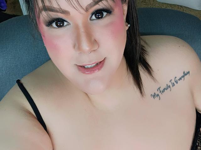 GiiannaMorrys with pink blush, black eyeliner, nose piercing, small tits, black bra, tattoo on right shoulder, smiling, brown hair, white skin, gray couch in background.