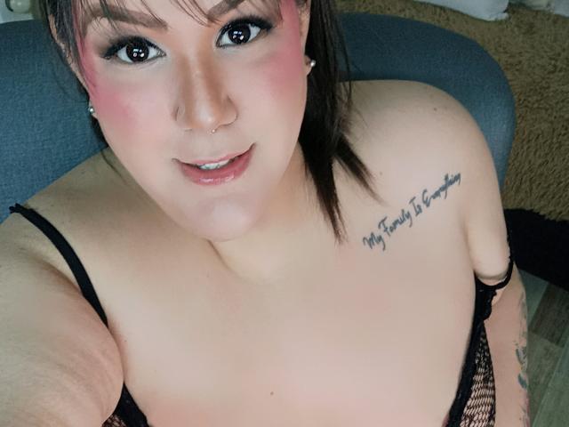 GiiannaMorrys with pink eye makeup, black eyeliner, nose ring, and tattoo on right shoulder, looking up, showing cleavage in black lingerie, smiling with slightly parted lips.
