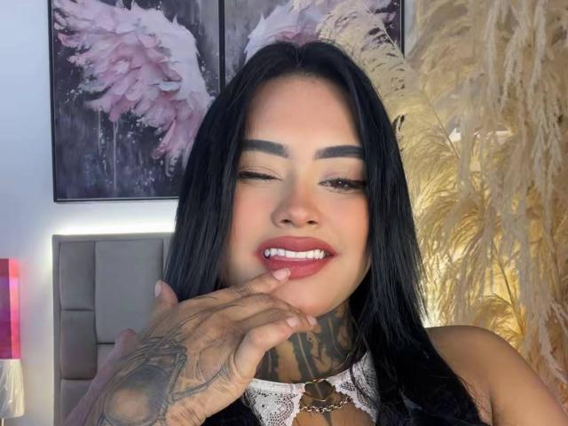 LairaHearvy with long black hair, winking, and biting her finger with tattoos on her hand, wearing lace and red lipstick, smiling with visible teeth.
