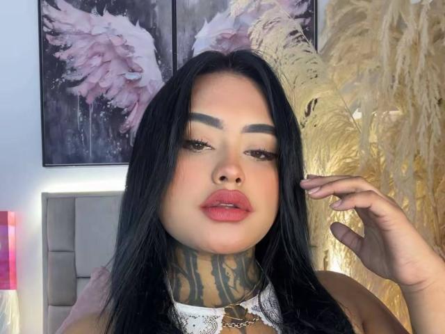 LairaHearvy with black hair, full red lips, and a tattooed neck, wearing a white lace top, touching her hair, with pink winged art in the background.