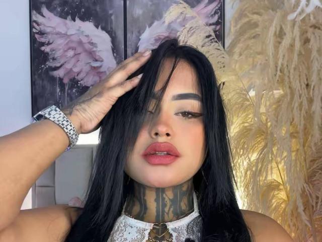 LairaHearvy has long black hair, tattoos on her neck, and is wearing a silver bracelet. She's touching her hair with one hand, showing pink lipstick and a sexy look.