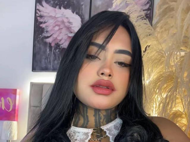 LairaHearvy has long black hair, thick eyebrows, pink lips, and tattooed neck with lace collar. Her makeup is bold with dark eyeliner. Her ass is not visible.