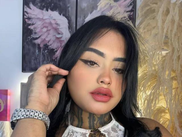 LairaHearvy with black hair, bold winged eyeliner, pink lips, white lace collar, tattooed neck, hand on head, silver bracelet, looking at camera, puffy pink lips, soft makeup.