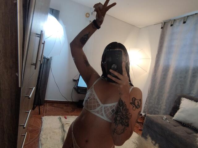 Mllevv in white mesh lingerie, showing off tattoos, making a peace sign, with wet hair, holding a phone in a mirror selfie. Her tits are small, her pussy is unseen.