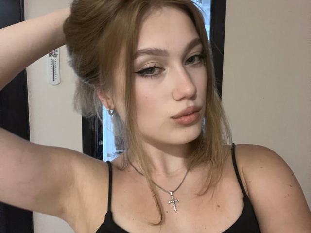 BobbyeWelson has light brown hair in a messy updo, wearing black spaghetti straps, a cross necklace, and eyeliner, with her arm raised, looking down with slightly parted lips.