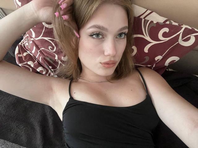 BobbyeWelson has light skin, green eyes, and pink nail polish, wearing a black tank top, lying on a bed with a red and white patterned pillow, looking at the camera.