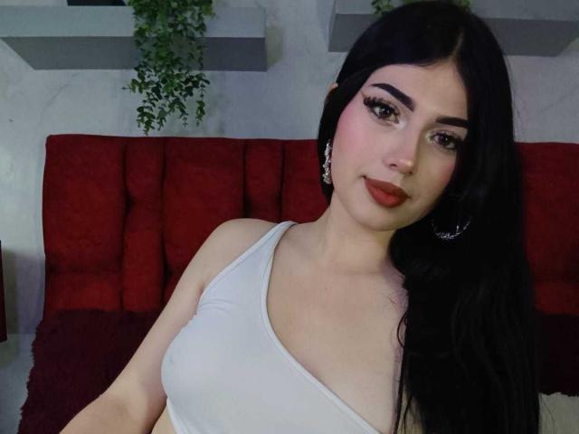 IsabellaaRossi with long black hair, white tank top showing her tits, red lipstick, and earrings, sitting on a red couch, looking at the camera with a slight smile.