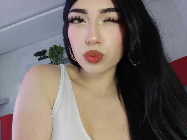 Isabella Rossi, met lang zwart haar, knipoogt, draagt een witte tanktop, rode lippenstift en gouden oorringen, en zet haar volle, rode lippen op een zuurmondje. Haar borsten zijn licht zichtbaar..