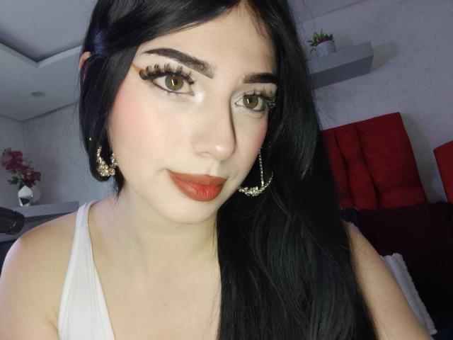 IsabellaaRossi has long black hair, green eyes, thick eyelashes, and red lipstick. She's wearing a white top and gold earrings, with a slight smile. Boobs not visible.