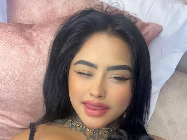LairaHearvy with winking eye, dark eyebrows, and pink lipstick, has black hair, a tattoo on her neck, and is wearing a chain necklace, looking sexy.