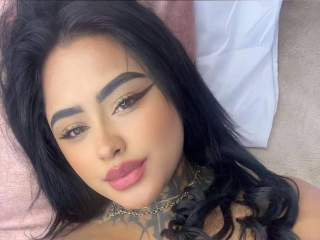 LairaHearvy has long black hair, thick black eyebrows, gold eyeliner, pink lipstick, and tattoos on her neck. She's wearing a chain necklace and lying on a pink pillow.