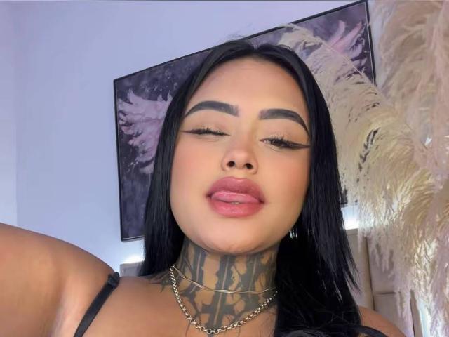LairaHearvy with black hair, tattoos on her neck, and a gold chain, has full pink lips and eyeliner, looking down with a slight smile. Boobs visible in black strap.