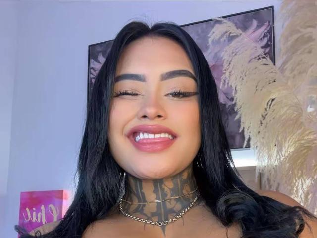 LairaHearvy has long black hair, tattooed neck, and is smiling with pink lipstick. Her eyes are closed, and she's wearing a chain necklace. Her tits are not visible.