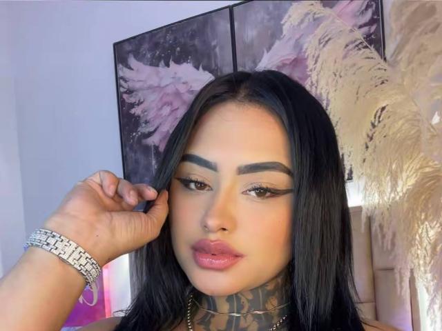 LairaHearvy has long black hair, thick eyebrows, winged eyeliner, pink lips, and a tattooed neck. She wears a silver bracelet and a gold necklace, looking at the camera.