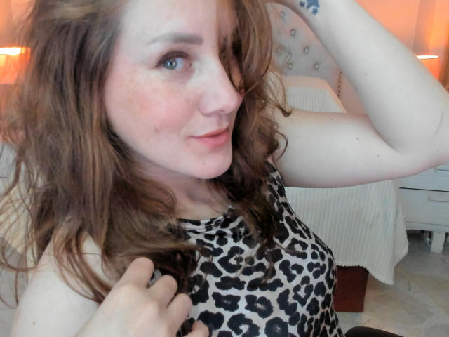 KatyaRogersX with wavy brown hair, wearing a leopard print top, holding her hair up, showing a small tattoo on her forearm, and smiling slightly.