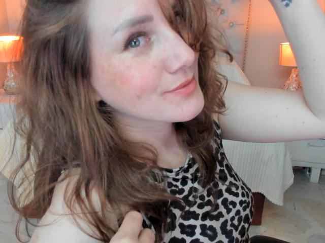 KatyaRogersX with wavy brown hair, wearing a leopard-print top, has fair skin and freckles, smiling with one hand in her hair, showing a small tattoo on her arm.