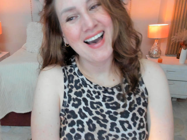 KatyaRogersX with smiling face, brown wavy hair, wearing leopard print sleeveless top, white bed with textured pillow, white nightstand with lamp, orange glow, white curtains in background.